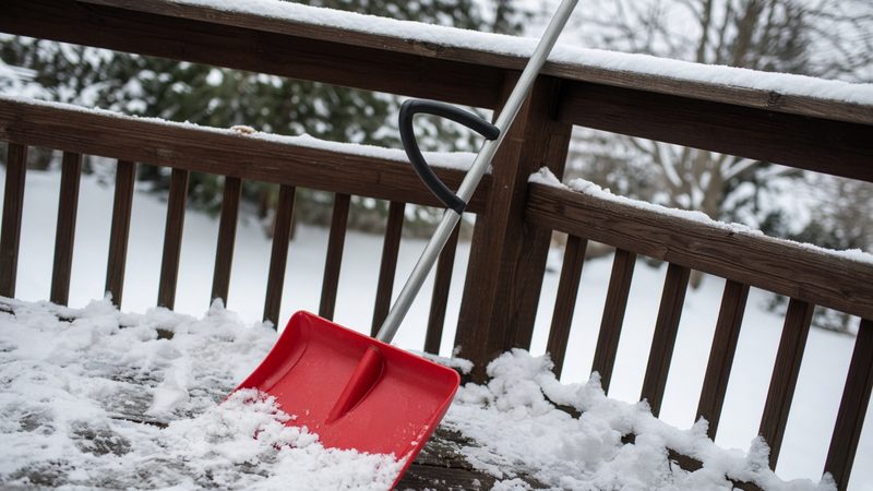 A red snow shovel with a second curved handle leaning on a snowy deck railing, in Pittsburgh, Pennsylvania